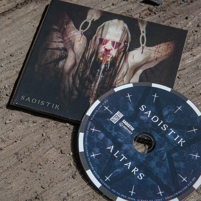 A Sadistik CD lies on a rugged wooden surface, its cover depicting a haunting figure with blood and chains. The disc features cross symbols, the album title, artist name, and glimpses of featured artists and the track list inside.
