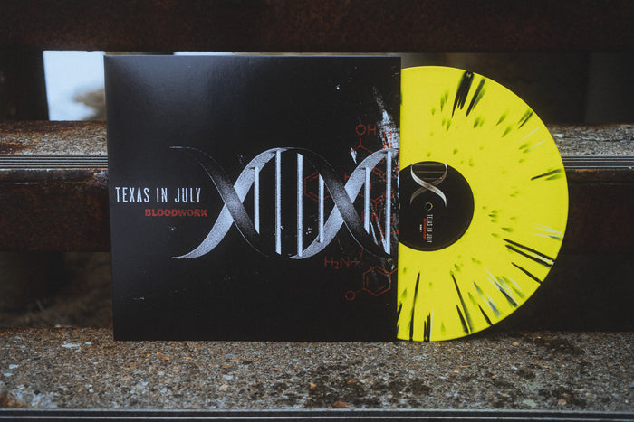The image displays the Texas In July album Bloodwork. Its cover has a DNA double helix with chemical formulas, and the vinyl features an eye-catching yellow splatter design accented by black streaks. The jacket and vinyl rest on rusty cement stairs.