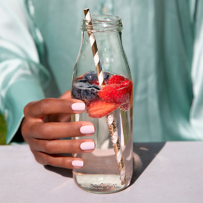 Wearing Manucurist's Blossom, a pale pink opaque creamy polish, a person holds a glass of water with a gold-striped straw, plus sliced strawberries and blueberries, their nails perfectly matching their light blue shirt.