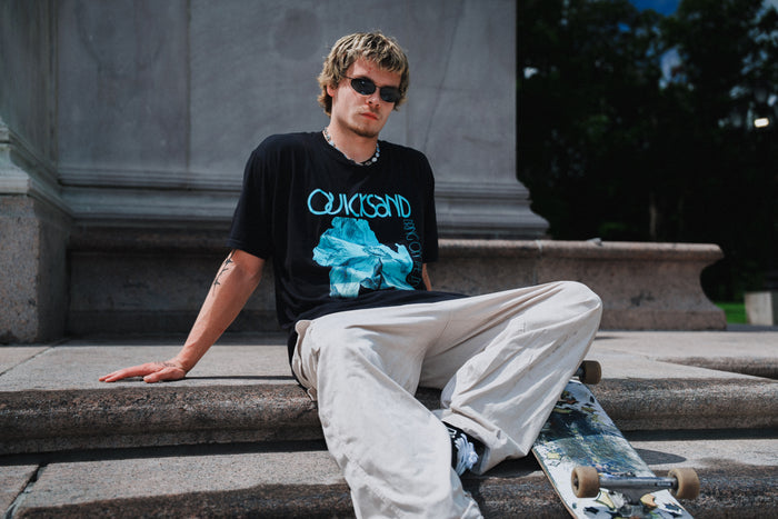 A person wearing sunglasses, beige pants, and a black Quicksand graphic t-shirt made from soft ring-spun cotton sits on stone steps beside a skateboard, outdoors near a large monument or building.
