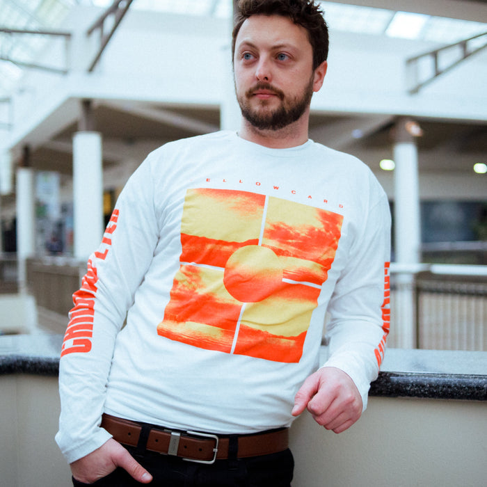 A man with short brown hair and a beard leans against an indoor railing, wearing a white Yellowcard long-sleeve T-shirt with an orange-yellow graphic design and text on the sleeves.