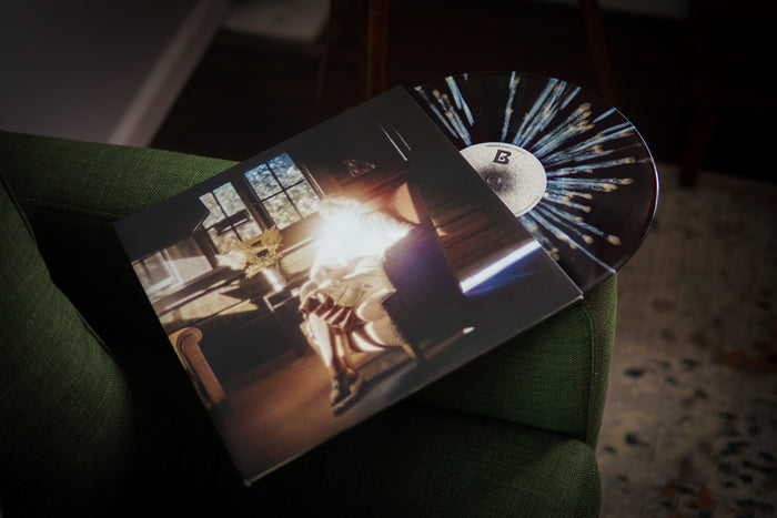 A transparent black vinyl with bone and white splatter peeks out of a jacket which shows a glowing, backlit figure in an armchair by a window in a sunlit rustic room. The vinyl and jacket rests on a green couch.