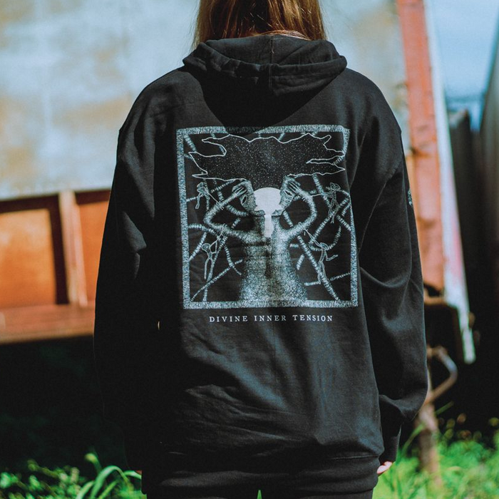 A person with long hair faces away, wearing a black Hail The Sun hoodie with an abstract tree design—roots and branches encircle a bright moon or sun. Below the image is the text “DIVINE INNER TENSION.”.