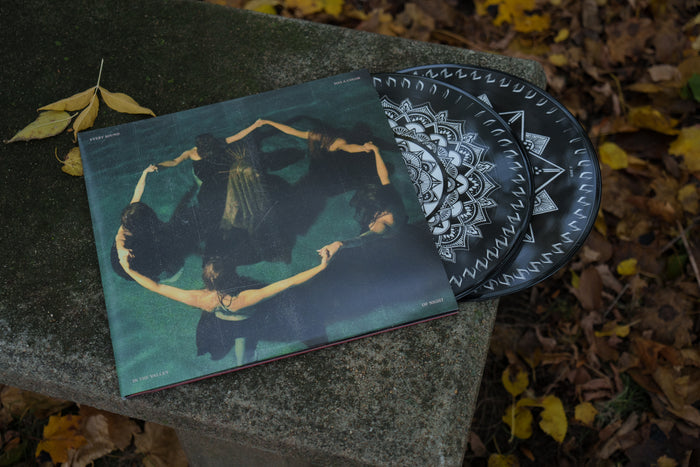 A Limited Edition Vinyl by Night Verses with a black and white geometric design partially slides out of its album cover, which shows women in black dresses holding hands in a circle, all resting on a stone bench amid fallen autumn leaves.