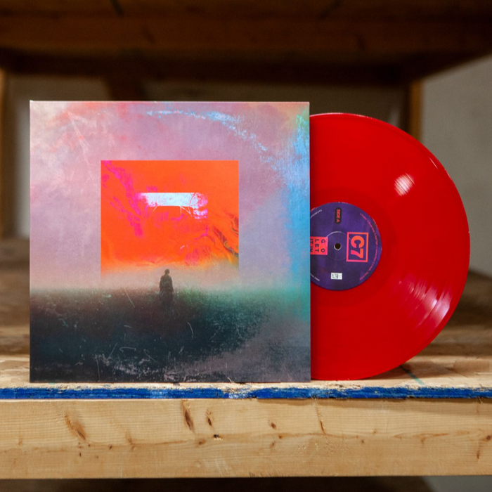Vinyl is Red on side A. The jacket is a hazy image of a man standing underneath a red square. The ground the man is standing on is black with a pink sky. The vinyl and jacket are propped against a wooden shelf.