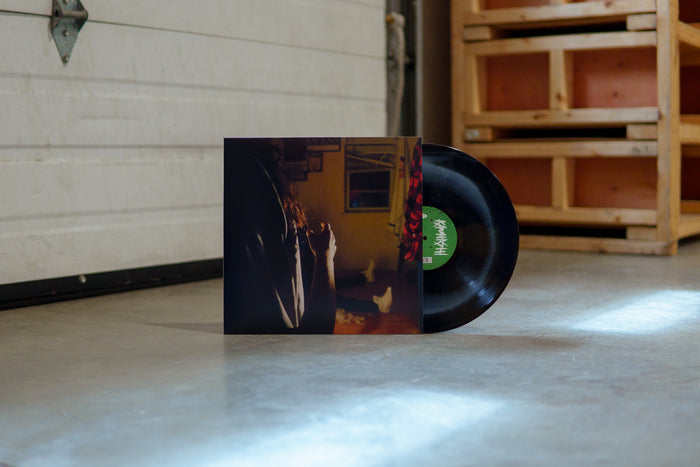A black vinyl with white crescent moon peeks out of a jacket with the image of three Kaonashi members in front of a window in a house with broken down blinds. The closest member is only showing his arms and hair, with the second only showing his red flannel, and the third sitting down with white shoes.