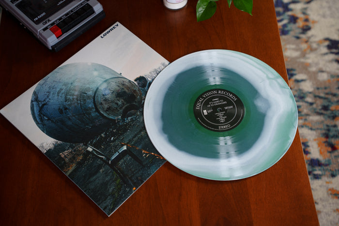 A limited edition green and white marbled vinyl of Lowrev’s album "It’s Hard to Lie to Strangers" rests partly on its cover, which shows a weathered metal tank outdoors. The Equal Vision Records label is visible on a wooden table.