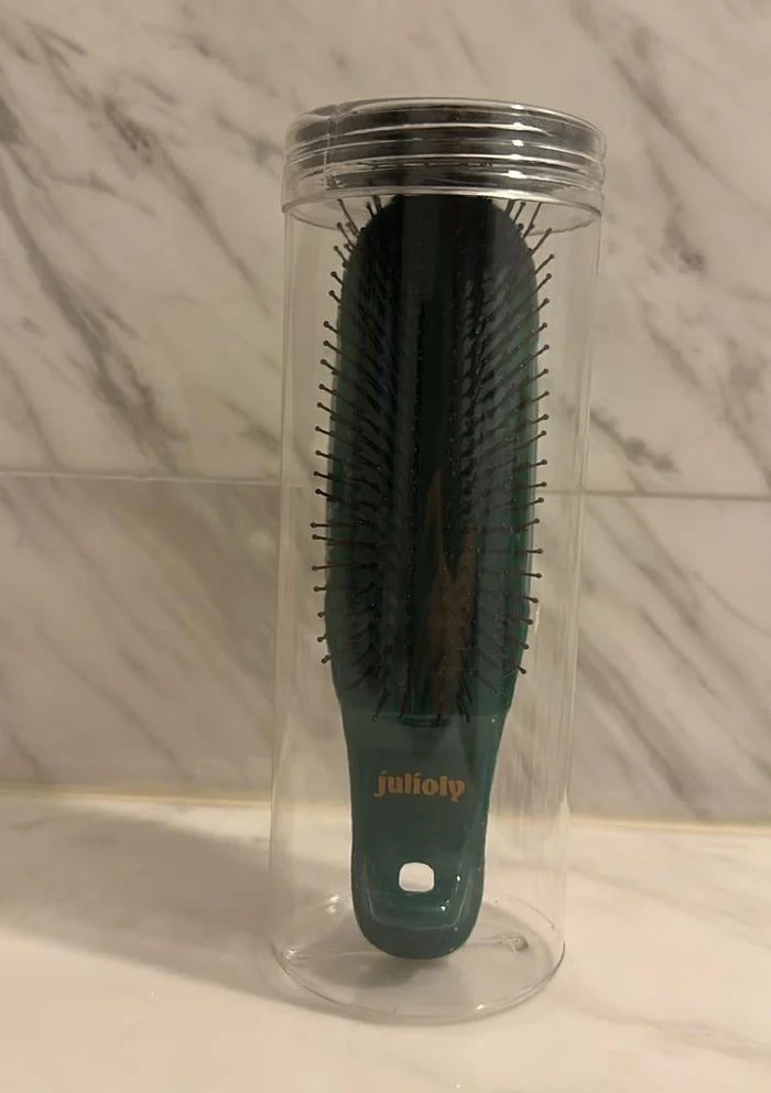 Black hairbrush with green handle in a clear plastic stand against a marble wall.
