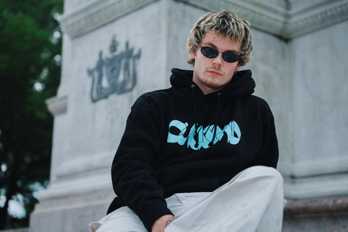 A young man wearing sunglasses, light pants, and a black Quicksand heavyweight pullover hoodie with a blue graphic and fleece-lined hood sits in front of a stone monument with engraved details and trees in the background.