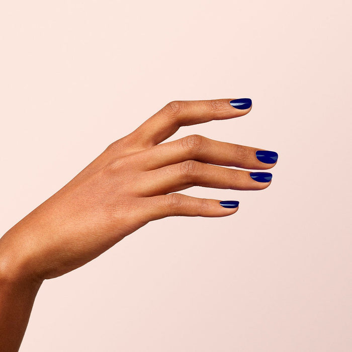 A hand with medium brown skin rests on a light beige background, nails neatly painted in Manucurist's Navy Blue gel polish and fingers gently curved in a relaxed pose.