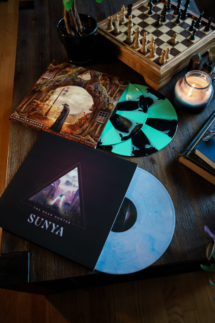 A wooden table holds a chessboard, a lit candle, a plant, and vibrant green-and-black patterned vinyl records—including The Dear Hunter's album Sunya—displayed near a scenic album cover.