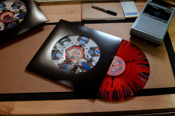 An Armor For Sleep red vinyl record with black streaks is partially pulled from a die-cut jacket with geometric art, displayed on a wooden desk next to a notepad, pen, and small tape recorder.