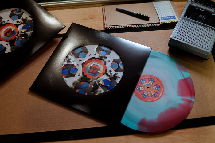 A colorful Armor For Sleep vinyl record with geometric artwork peeks out of a die-cut jacket, resting on a desk beside a notebook, pen, cassette tape, and portable tape recorder.