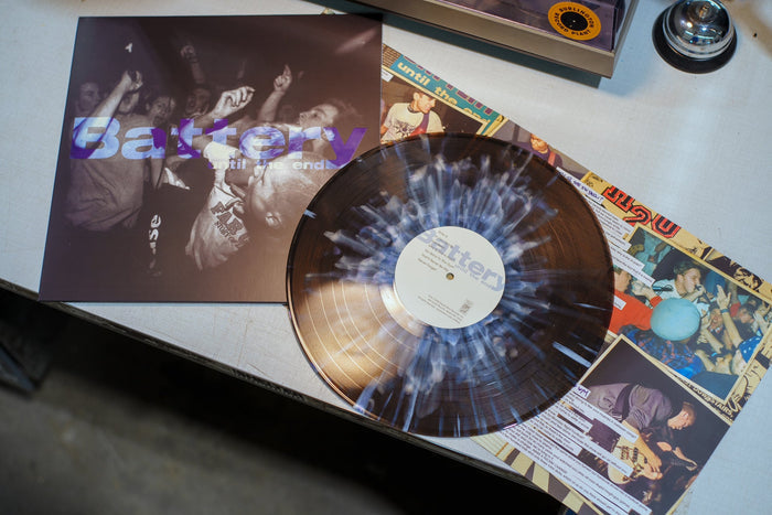 A translucent, limited edition Battery vinyl record with a white and blue splatter pattern and its cover—featuring a black-and-white concert crowd photo—rest on a table.