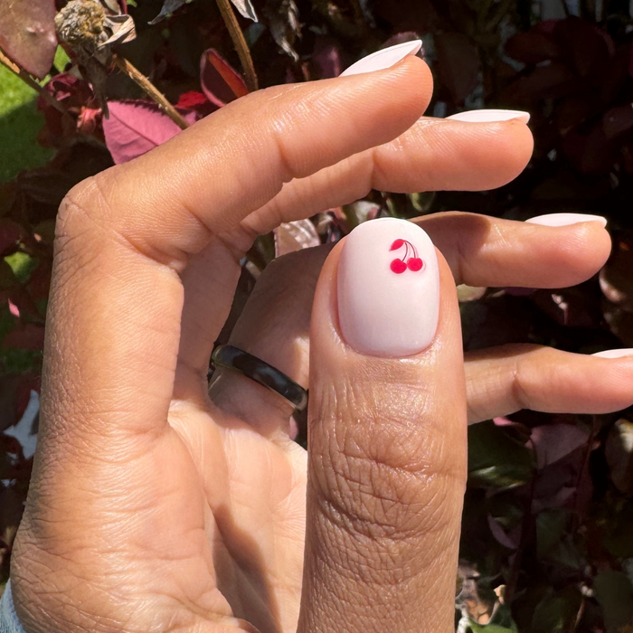 Vampy Cherry | Extra Short | Round - Tab Press-On nails with pastel pink base and red cherry detail on thumb in natural light outdoors