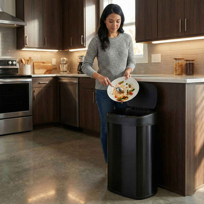 A woman in a modern kitchen using the iTouchless ITOS13BK black stainless steel 50L sensor trash can to throw garbage away.