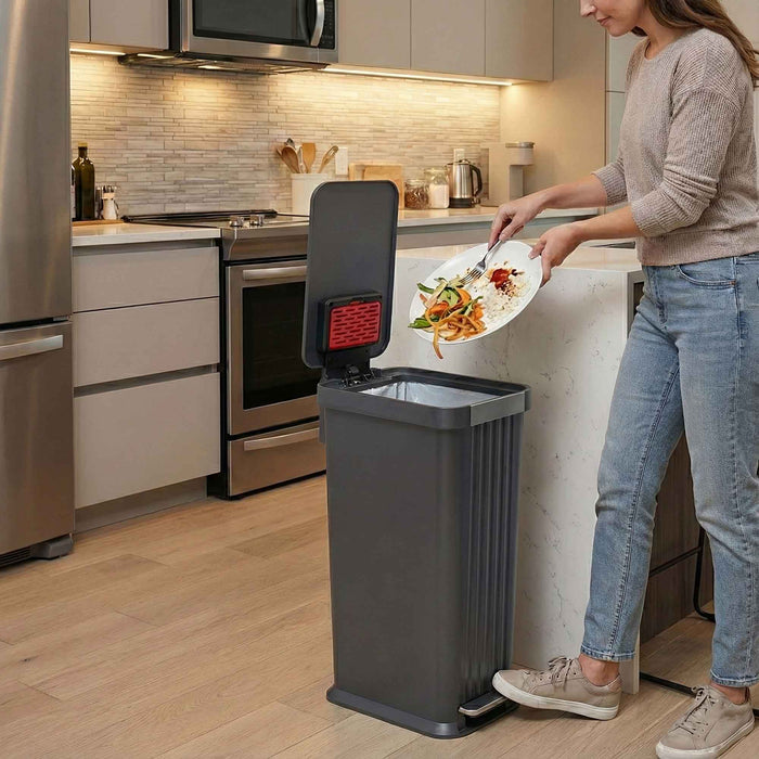 A woman in a modern kitchen using the iTouchless PE13GG gray step pedal trash can 50L to scrape food scraps off a plate.