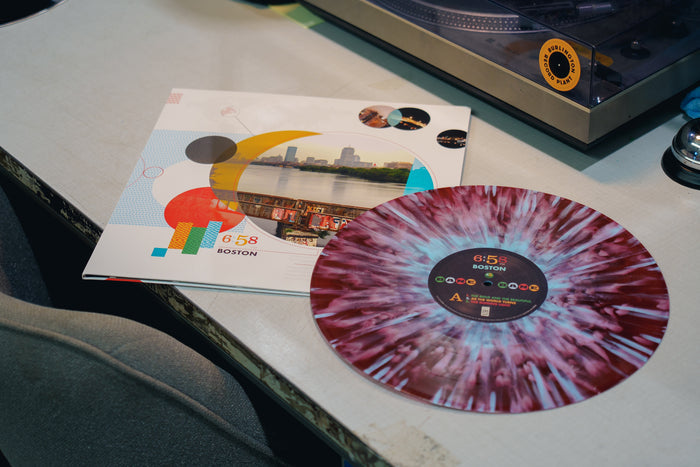 A marbled purple and white “6:58 Boston” vinyl record by Bane rests next to its vibrant sleeve—featuring a city skyline, geometric designs, and bold text—on a desk with a turntable nearby, evoking The Young and the Restless logo.