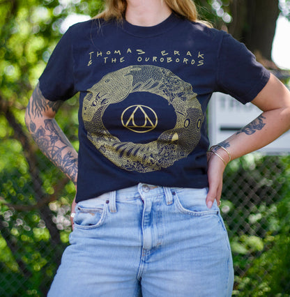 A person stands outside wearing a black Thomas Erak & The Ouroboros T-shirt featuring a gold ouroboros snake design and light blue jeans. Their tattooed arms are visible, with green foliage blurred in the background.