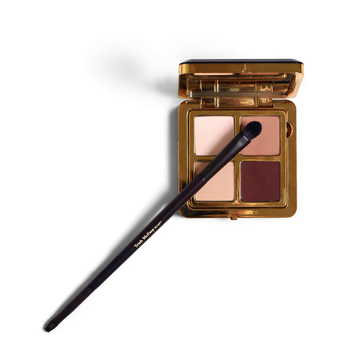 A four-shade eyeshadow palette in a gold compact with a black applicator brush resting across it, photographed on a clean white background.