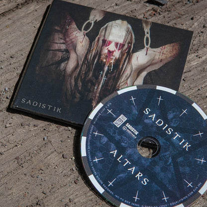 A Sadistik CD lies on a rugged wooden surface, its cover depicting a haunting figure with blood and chains. The disc features cross symbols, the album title, artist name, and glimpses of featured artists and the track list inside.