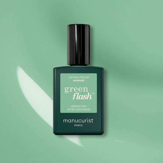 A bottle of Manucurist Amande Green Flash nail polish, featuring a black cap, is shown on a matching green background with light reflecting beside it.