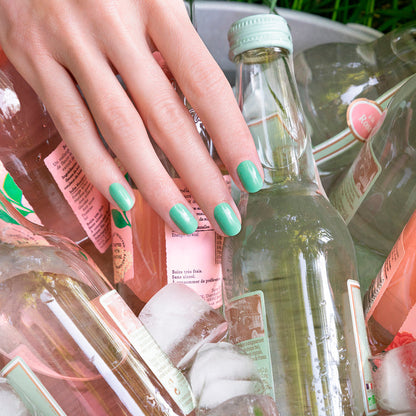 A hand with Manucurist's Amande green nail polish rests on glass bottles of pink and clear drinks, surrounded by ice cubes in a metal bucket.