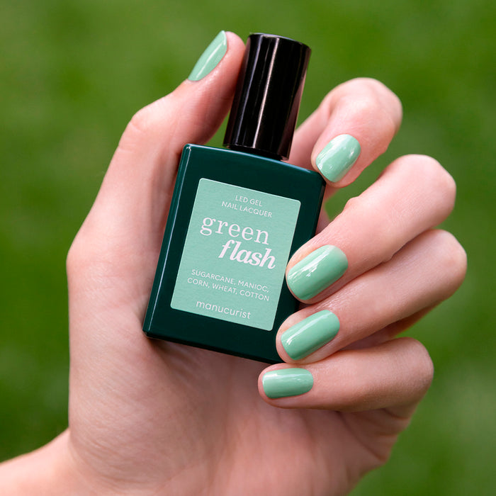 A hand with neatly manicured, mint green gel polish nails holds a bottle of Manucurist Amande LED gel nail lacquer against a blurred green background.
