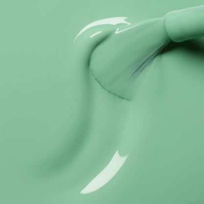 A close-up of a brush applying Manucurist’s Amande gel polish in smooth mint green, with glossy, reflective streaks visible in the fresh paint.