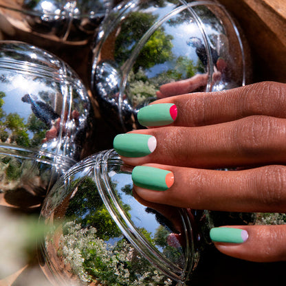 A hand with modern nail art showcases Manucurist's Amande, an almond green polish, with multicolored half-moon tips. The nails rest on shiny metallic balls reflecting trees and sky.