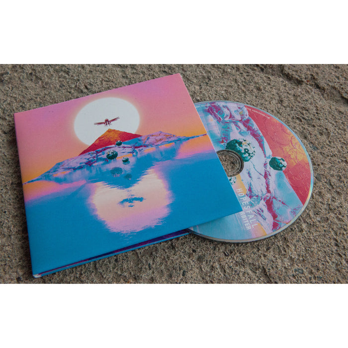 Square CD with artwork that is the sun with a bird with it's wings spread over it. Below the bird is a mountain in the center of a body of water. The sun is setting and there is a reflection in the water of the mountain, sun, and bird. CD is peeking out of CD cover and on a brown surface.