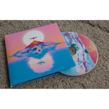 Square CD with artwork that is the sun with a bird with it's wings spread over it. Below the bird is a mountain in the center of a body of water. The sun is setting and there is a reflection in the water of the mountain, sun, and bird. CD is peeking out of CD cover and on a brown surface.