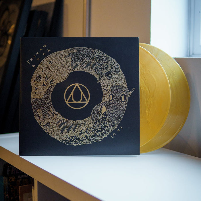 A Limited Edition Gold Nugget LP by Thomas Erak & The Ouroboros, with yellow transparent discs and a black sleeve with intricate gold artwork and geometric logo, sits on a white shelf near a window.