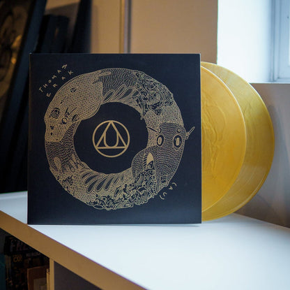 A Limited Edition Gold Nugget LP by Thomas Erak & The Ouroboros, with yellow transparent discs and a black sleeve with intricate gold artwork and geometric logo, sits on a white shelf near a window.