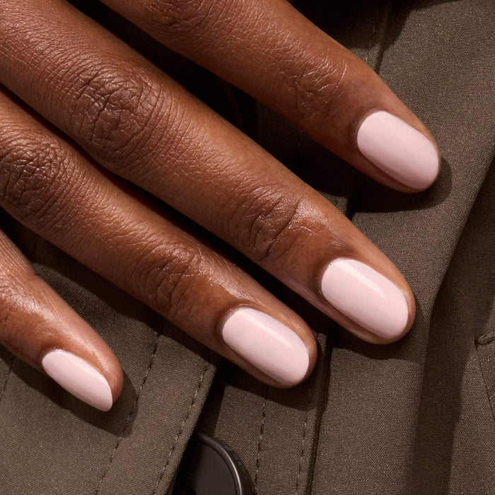 A close-up of a hand with short, manicured nails painted in Manucurist's Blossom, a pale pink gel polish, resting on brown fabric with visible stitching and a black button.