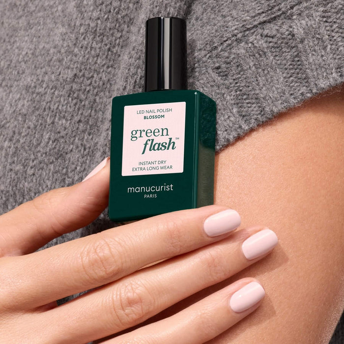A hand with light pink polished nails holds a green Manucurist bottle in the shade Blossom against a gray textured fabric background.