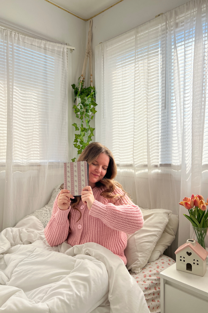 Woman in a pink sweater reading a kindle in a cozy bedroom with white curtains and a small plant.