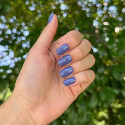 Hand showcasing Bootcut | Short | Squoval - Tab Press-On nails in glossy denim blue polish outdoors with green foliage background