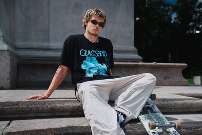 A person wearing sunglasses, beige pants, and a black Quicksand graphic t-shirt made from soft ring-spun cotton sits on stone steps beside a skateboard, outdoors near a large monument or building.