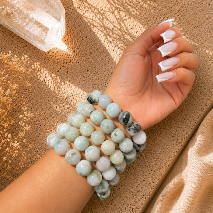 A close-up of a person's wrist wearing multiple beaded Jade bracelets made of pale green, white, and gray marbled stone, photographed on a textured tan surface with warm sunlight and shadows.