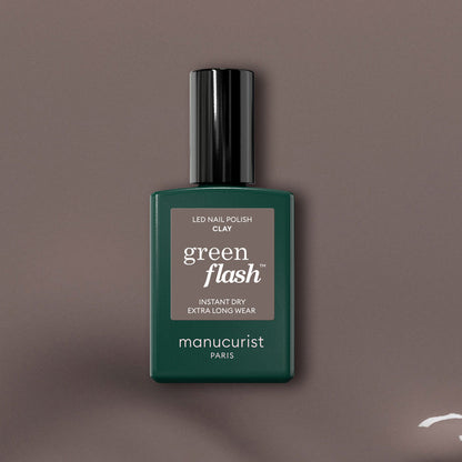 A bottle of Manucurist Clay Green Flash gel polish is centered on a taupe background, showcasing the dark green bottle with black cap and white text that highlights this innovative taupe shade.