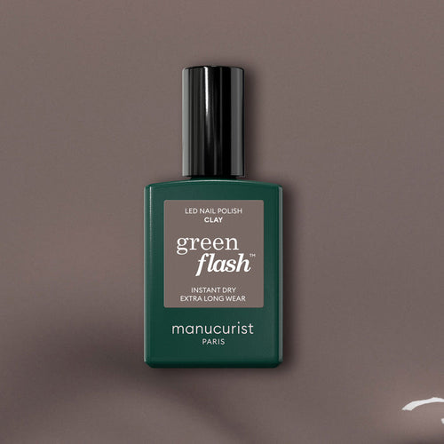 A bottle of Manucurist Clay Green Flash gel polish is centered on a taupe background, showcasing the dark green bottle with black cap and white text that highlights this innovative taupe shade.