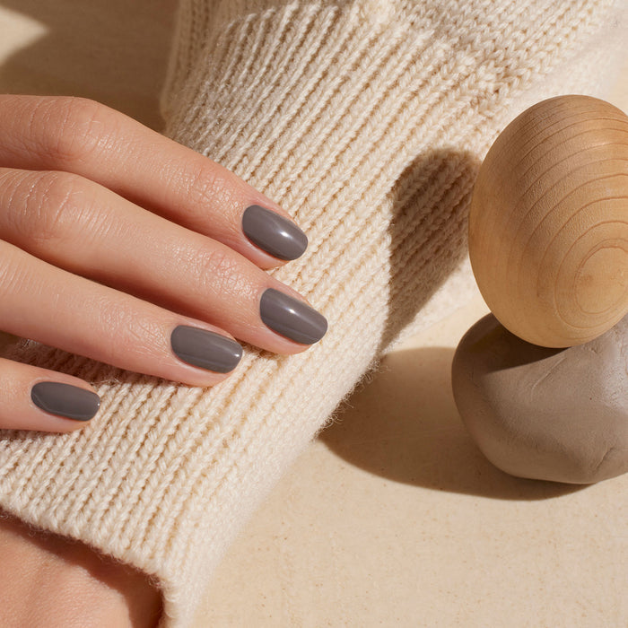 A hand with neatly manicured nails painted in Manucurist Clay rests on a cream knit sweater beside a wooden sphere and a piece of clay.