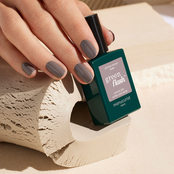 A hand with neatly manicured, chestnut gray nails holds a green bottle of Manucurist Clay polish against a beige, textured background.