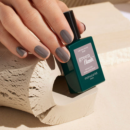 A hand with neatly manicured, chestnut gray nails holds a green bottle of Manucurist Clay polish against a beige, textured background.