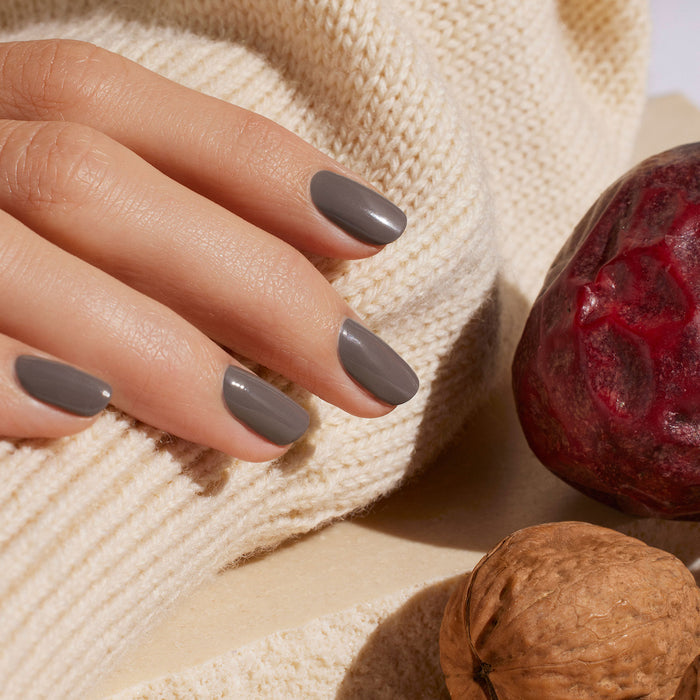 A hand with neatly manicured Manucurist Clay nail polish rests on a cream-colored knitted sweater, next to a red plum and a walnut.
