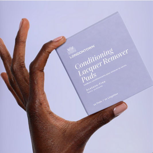 A hand displaying a box of LONDONTOWN Conditioning Lacquer Remover Pads with perfectly manicured nails against a soft lavender background—ideal for easy, on-the-go nail polish removal.