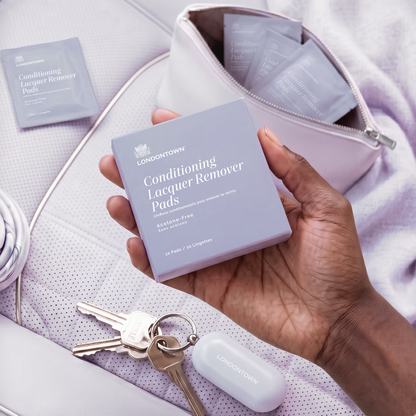 A hand holds a box of LONDONTOWN Conditioning Lacquer Remover Pads above a purse, showcasing the ideal travel-friendly nail polish removal option, with additional packets, a keychain, and a LONDONTOWN case visible nearby.