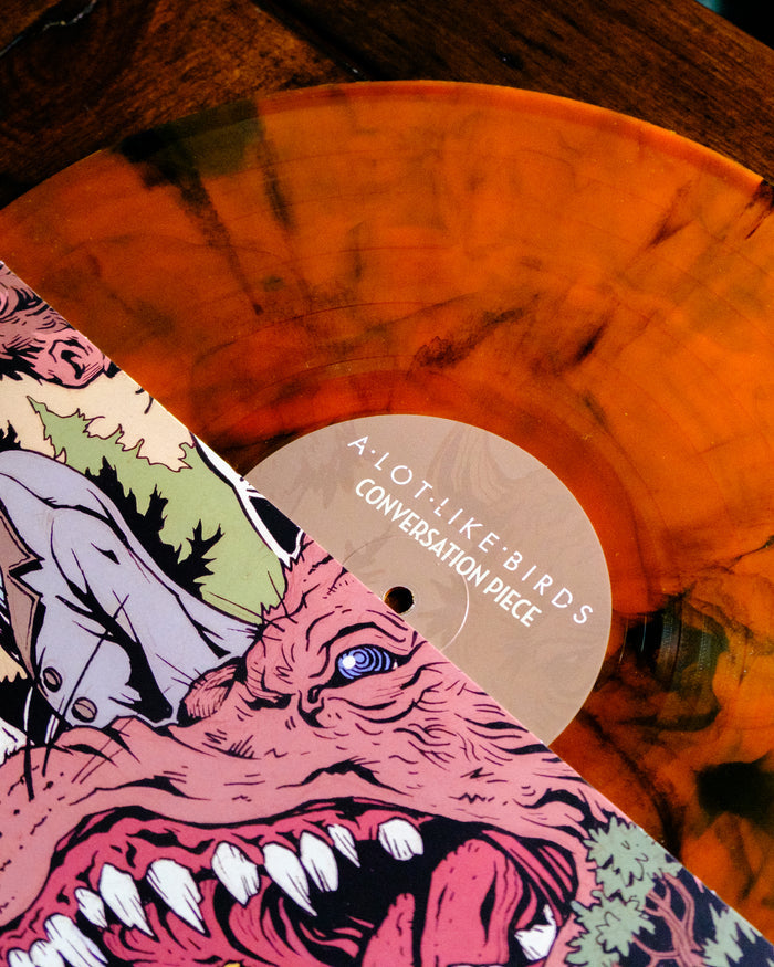 A Limited Edition orange vinyl with black marbling from A Lot Like Birds is partially covered by a sleeve featuring colorful, abstract snarling faces art and labeled "Conversation Piece.