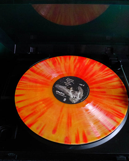 A bright orange and yellow Sparta "Marigold" vinyl record spins on a black turntable. This limited edition release features a center label with white text and an astronaut illustration, while the reflective turntable lid is partially open above.